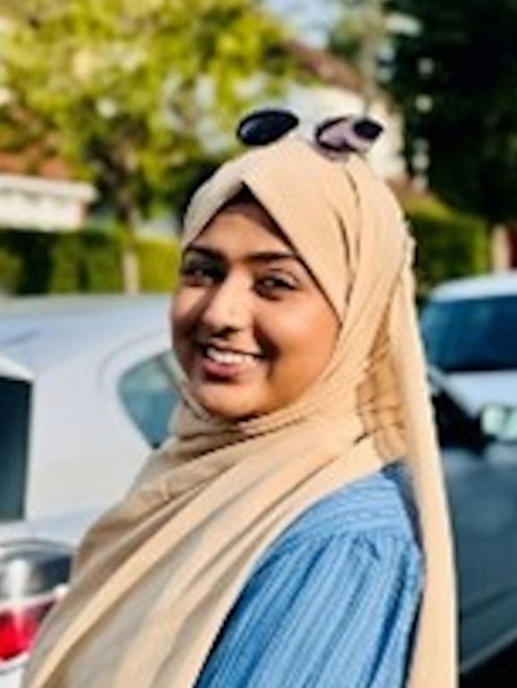 A person wearing a beige hijab and blue top smiles outdoors, with sunglasses resting atop their head.